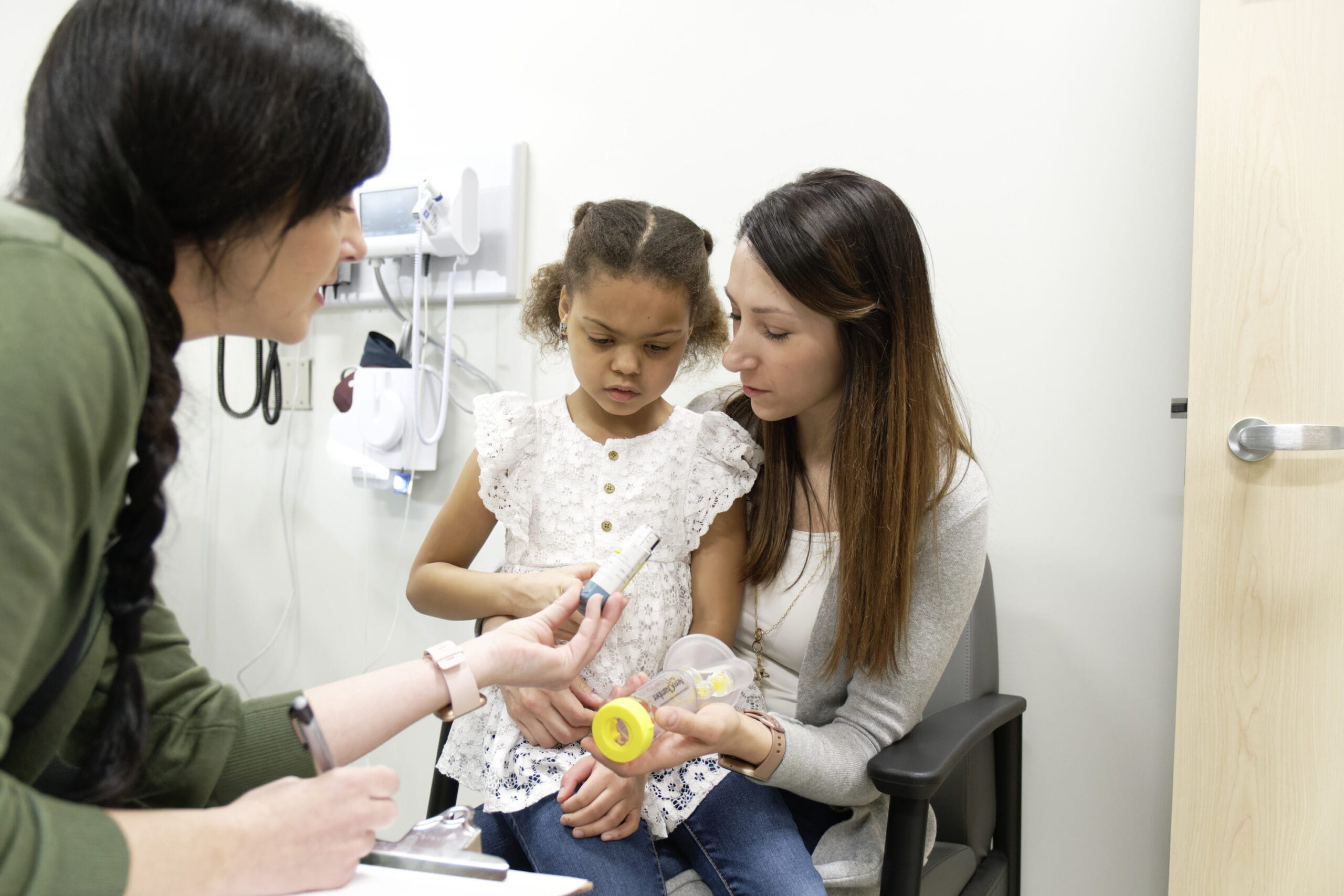 female nurse practitioner handing an inhaler to a female child sitting on her mother's knee.